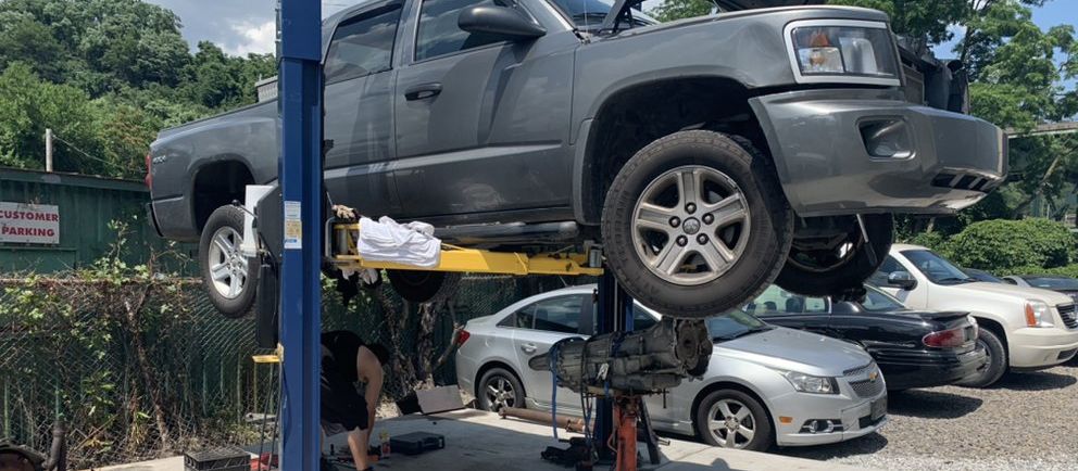 A grey pickup raised on a two-post lift in the yard with its hood up, other cars parked beyond it.